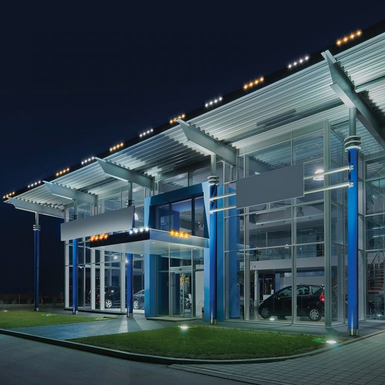 Car dealership showroom with white and blue lighting along the canopy at night.