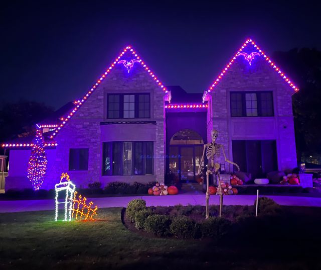 Stone two-story home lit in purple for Halloween, with lit bat shapes at the gable peaks and a skeleton in the yard.