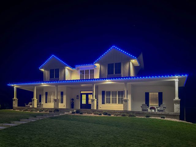White two-story farmhouse with a wide front porch, blue permanent lighting along the roofline and porch at night.
