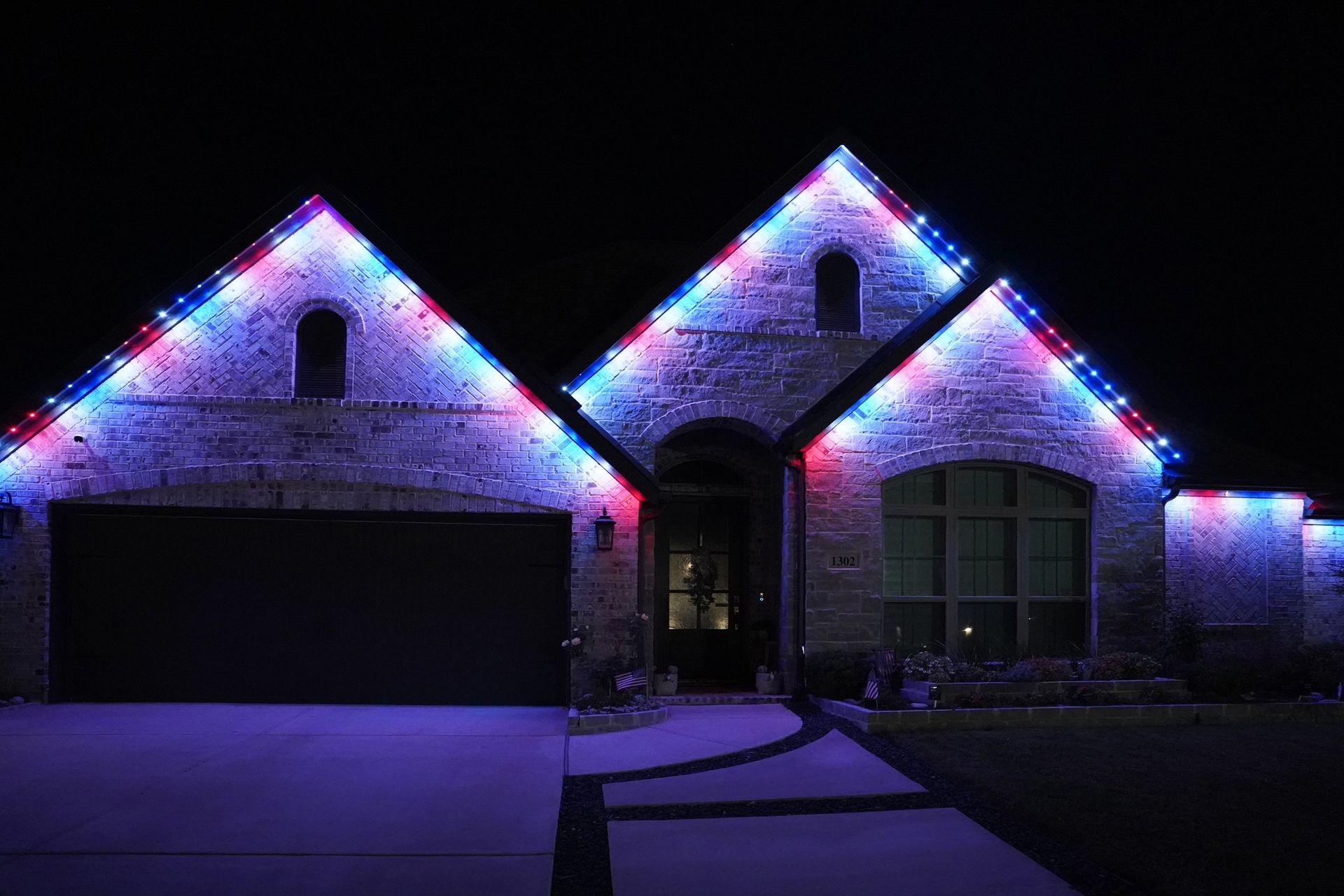 Stone and brick home at night with red, white and blue permanent lights along three front gables, washing the stone in purple.