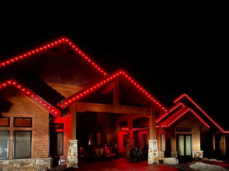 Stone and wood-sided home with several gables lit in red lights along the roofline at night.