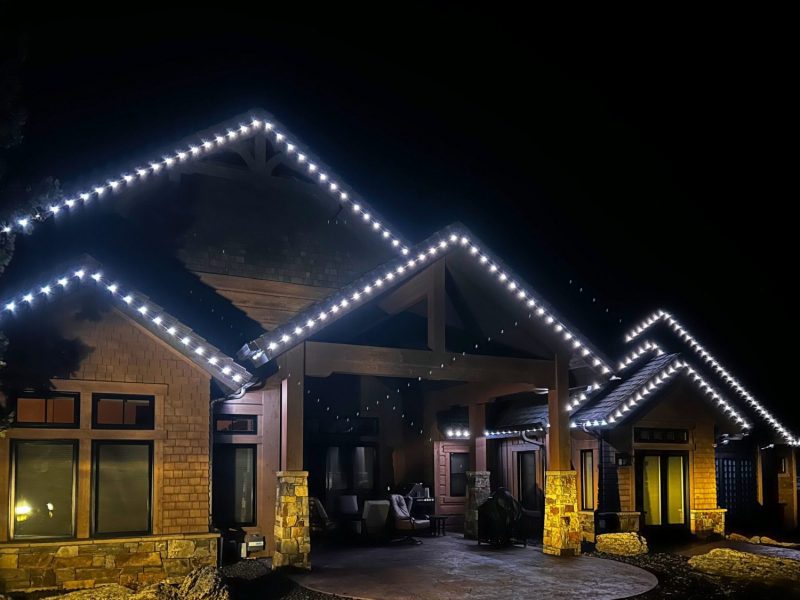 Stone and wood-sided home with multiple gables lit in white lights along the roofline, covered patio below.