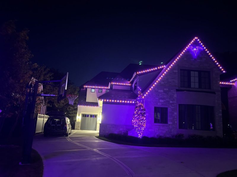 Stone two-story home lit in purple and orange for Halloween, with a lit bat shape at the roof peak.