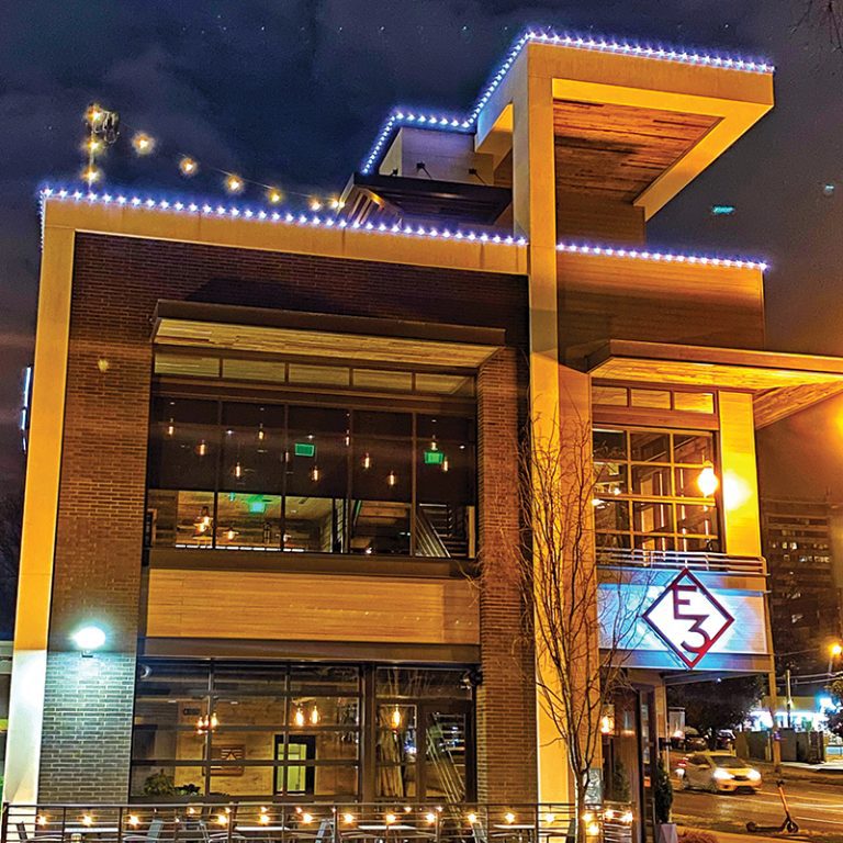 Multi-story building with lighting along the roofline, outdoor patio seating below.