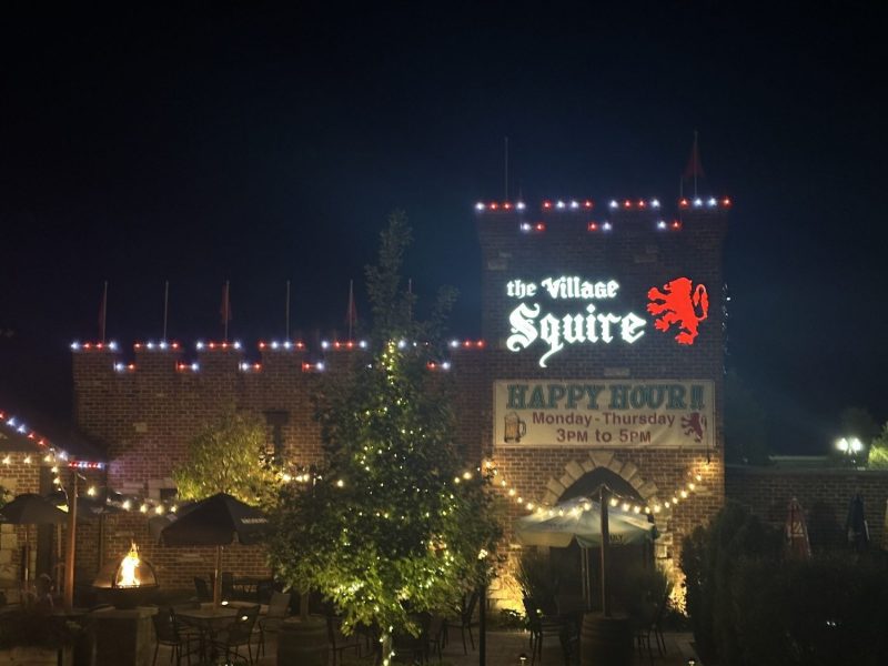 The Village Squire restaurant patio lit with string lighting at night, with outdoor seating and umbrellas.